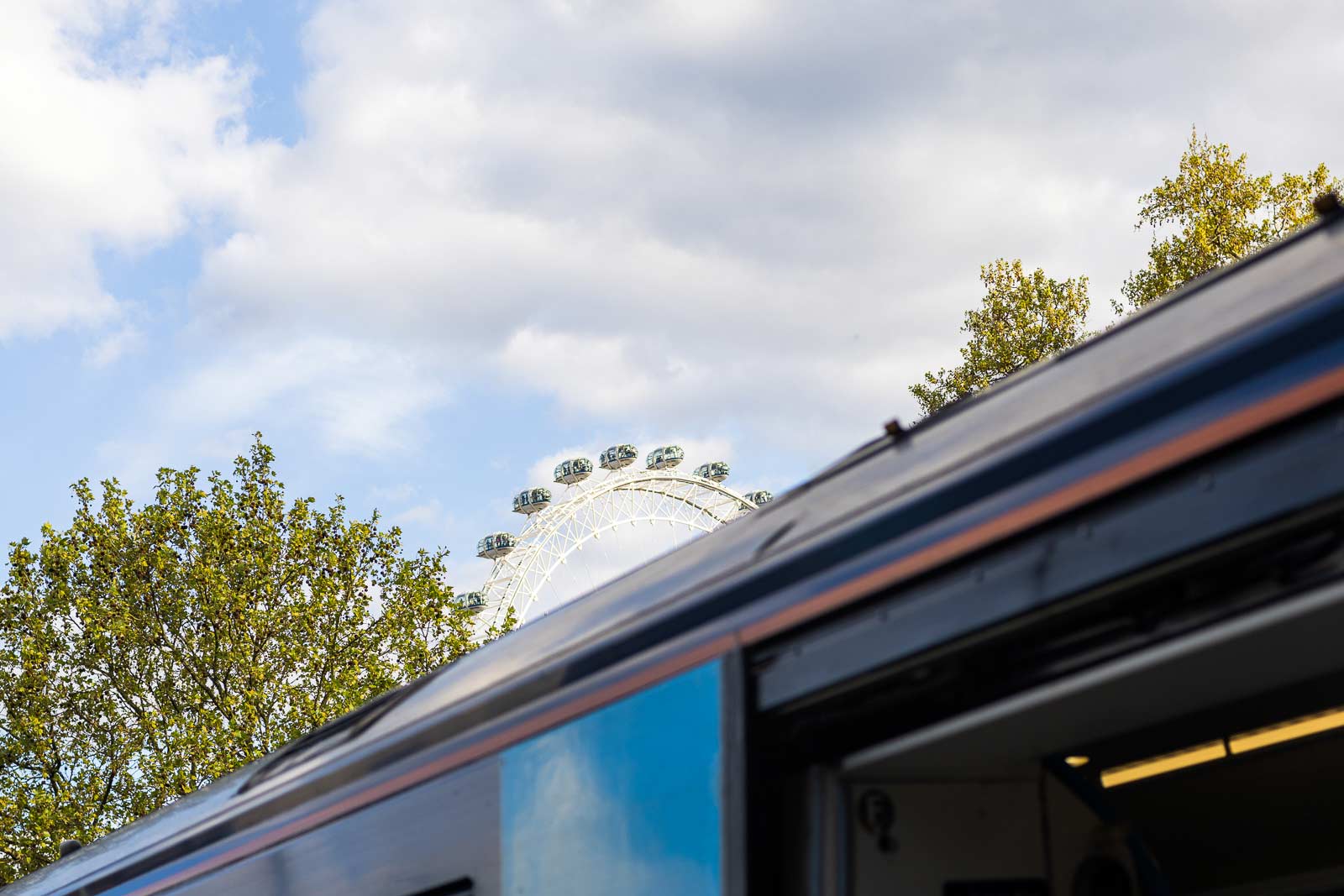 View of the London Eye behind a partially open train with trees and cloudy sky.