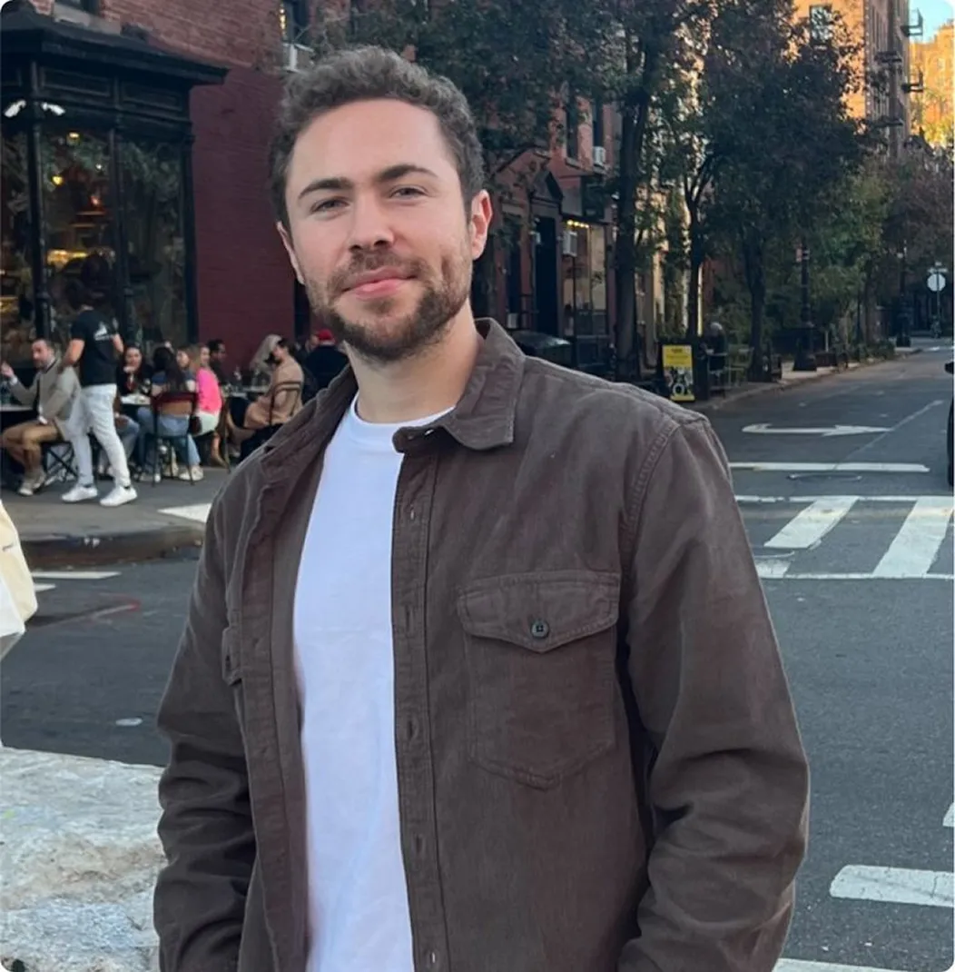 Young man with beard wearing a white shirt and brown jacket standing on a city street.