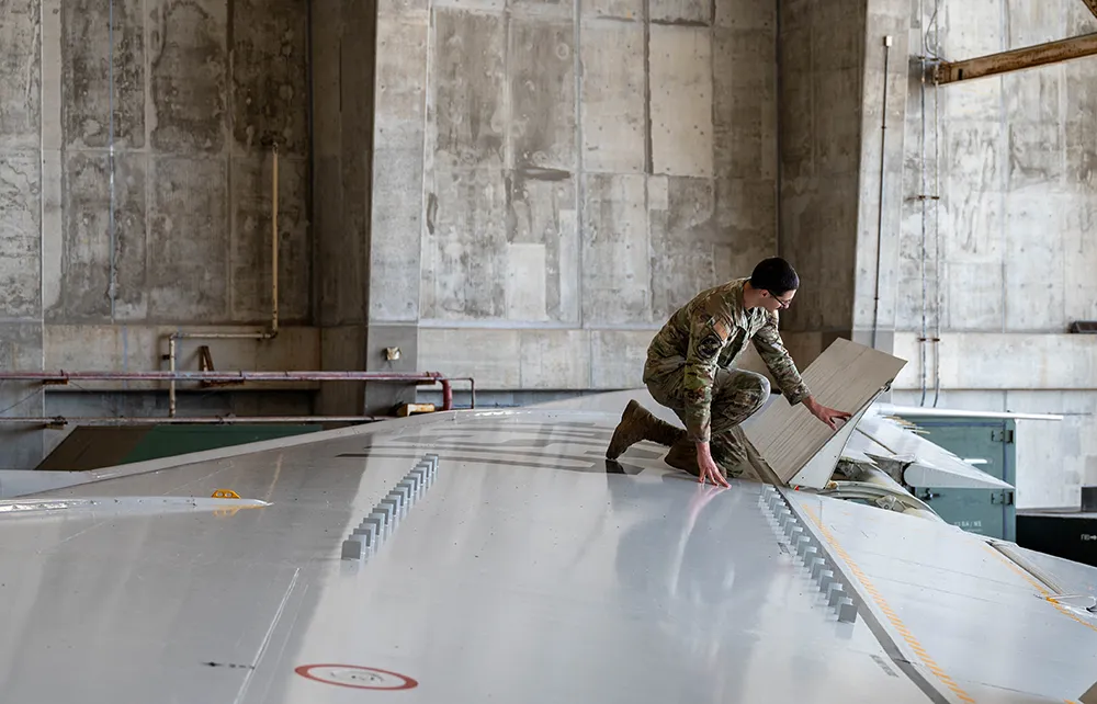 Military personnel in uniform inspecting or repairing a panel on an aircraft wing inside a hangar.