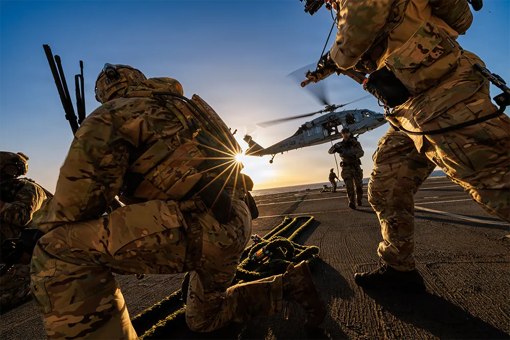 Soldiers in camouflage gear on a deck with a helicopter hovering at sunset.