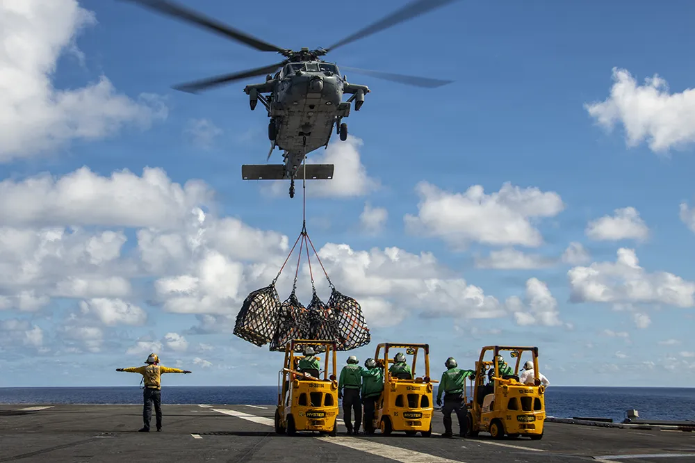 Helicopter lifting cargo nets above a ship deck with workers and forklifts below.