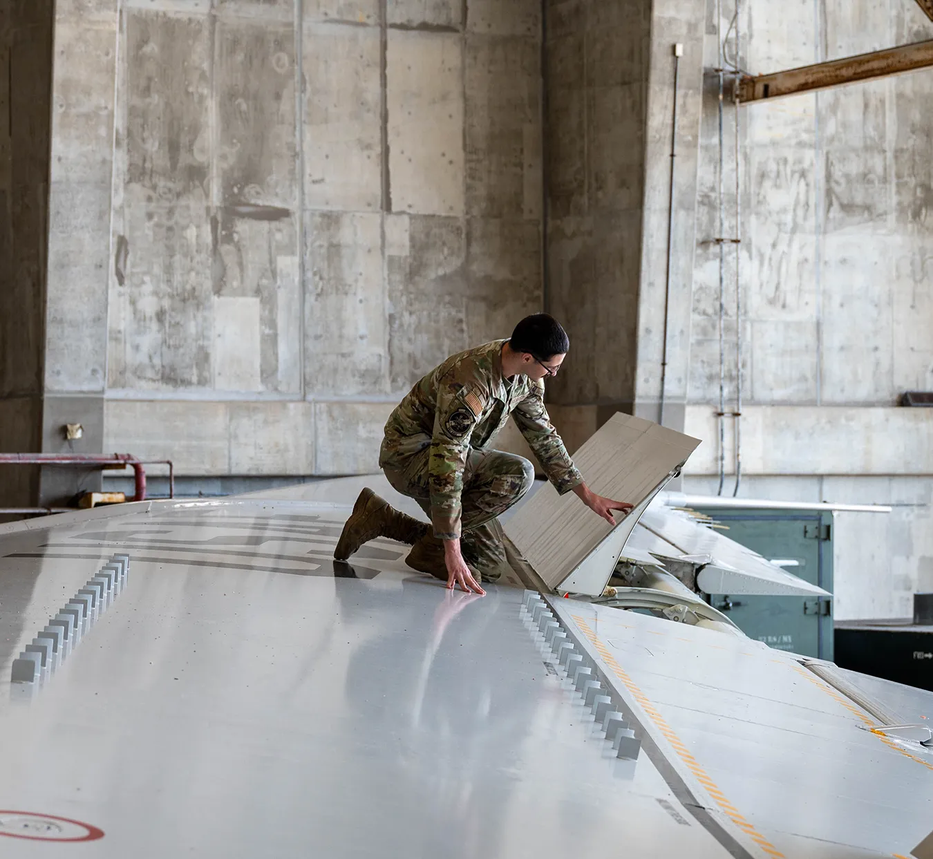 Military servicemember in camouflage kneels on an aircraft wing inspecting an open panel.