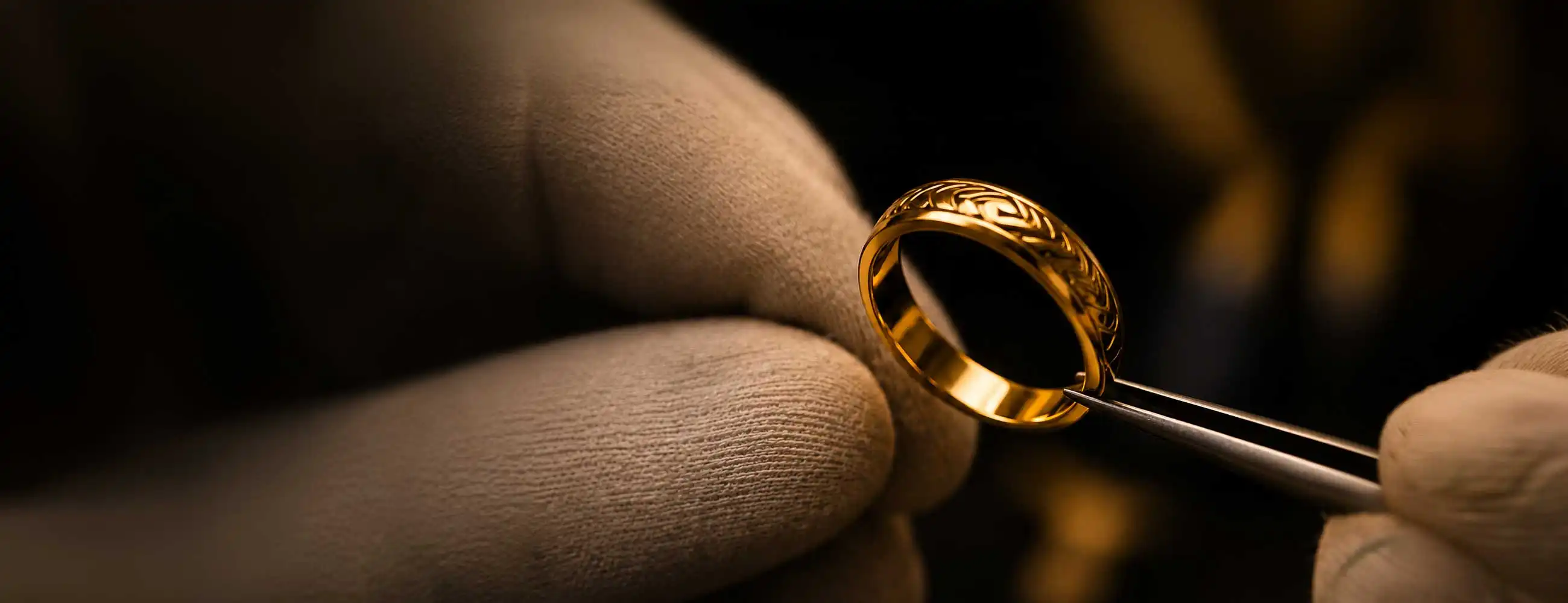 Close-up of an engraved gold ring held in tweezers against beige gloved fingers, lit warmly against a dark blurred background.