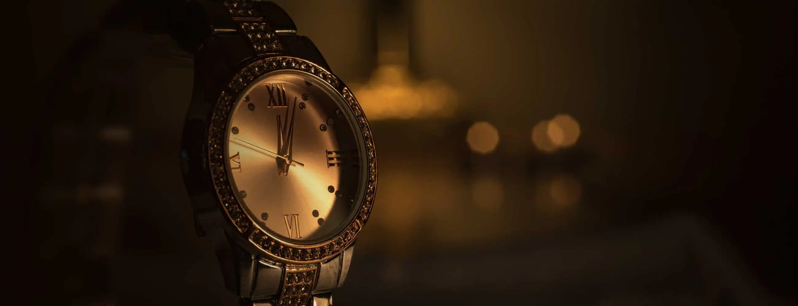 Luxury gold wristwatch with Roman numerals and crystal bezel, illuminated by warm ambient lighting against a blurred elegant background.