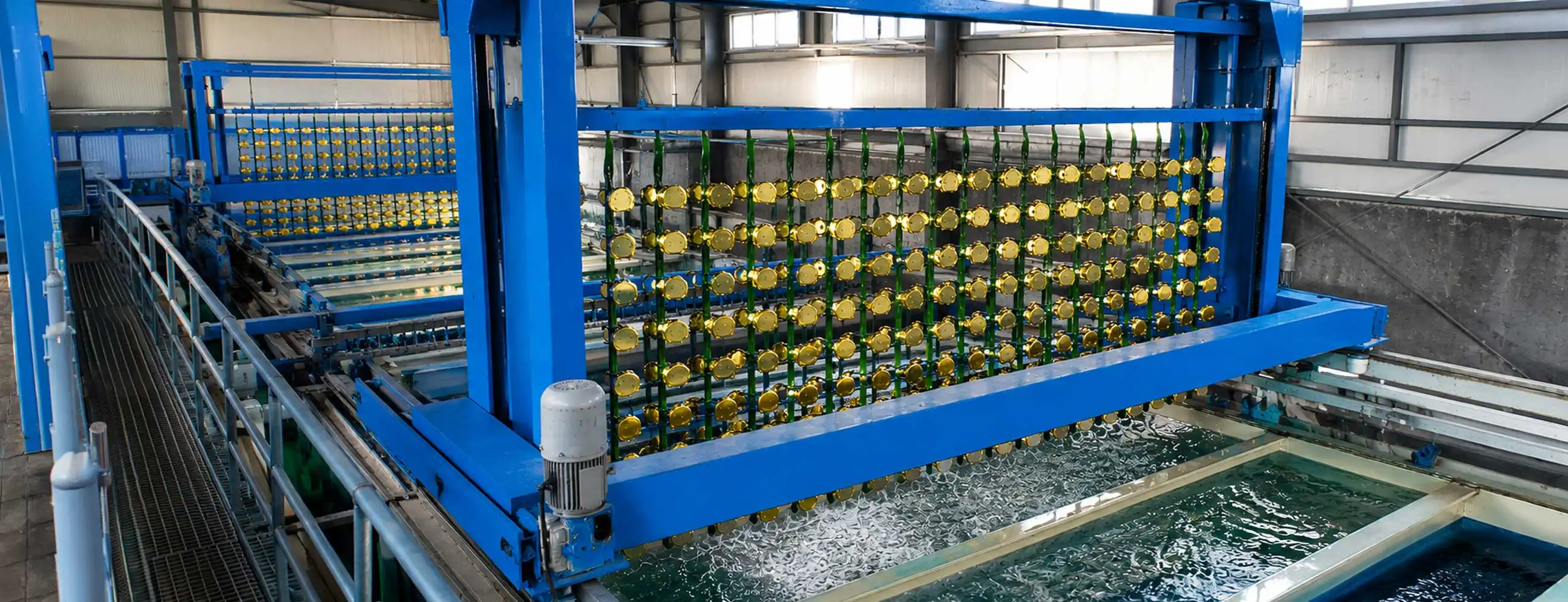 High-resolution industrial electroplating facility with blue metal gantry, black valves, and rows of gold-coated components suspended above bubbling chemical tanks in a modern factory setting.