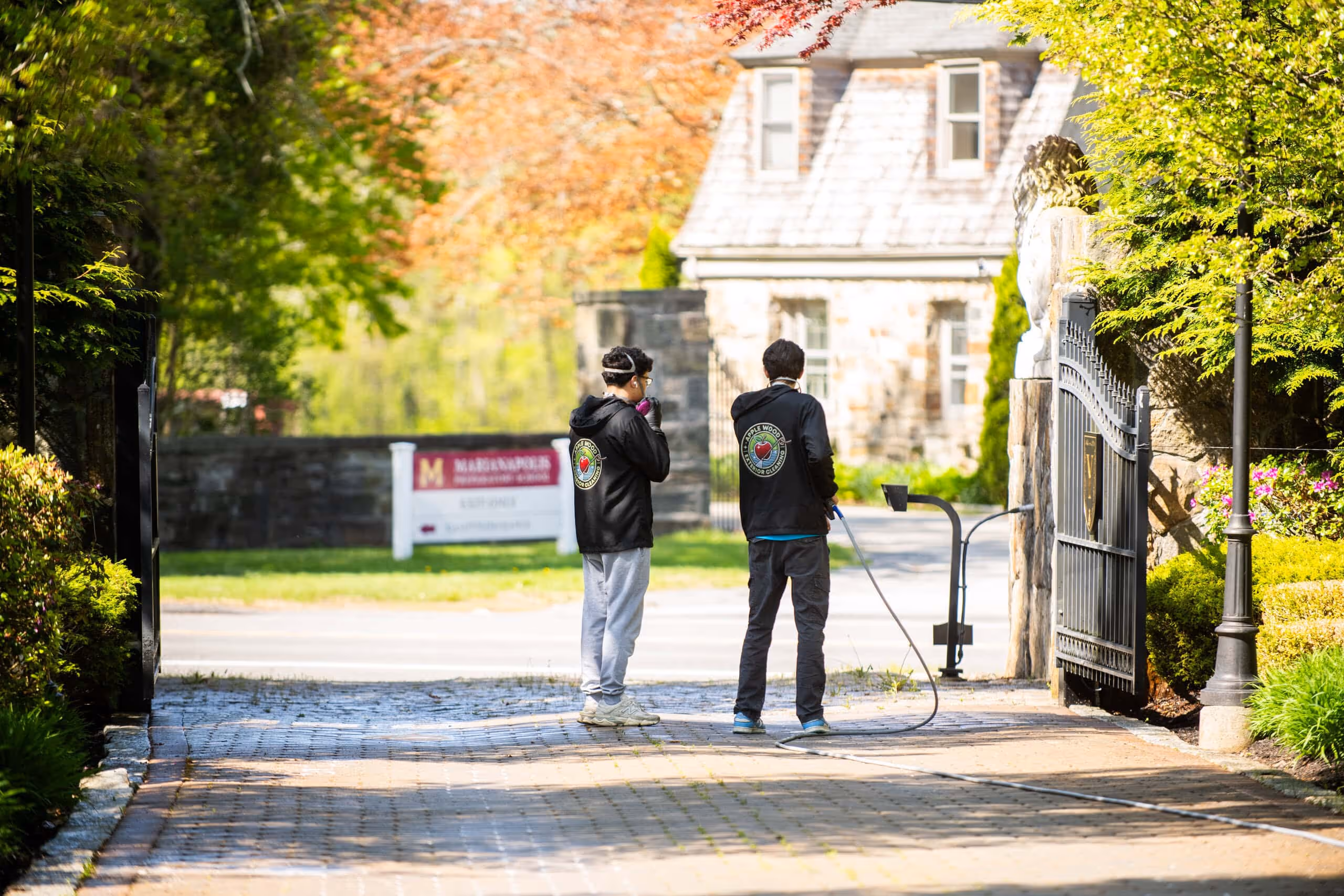 Two workers in black hoodies pressure washing a stone driveway at a gated property entrance.