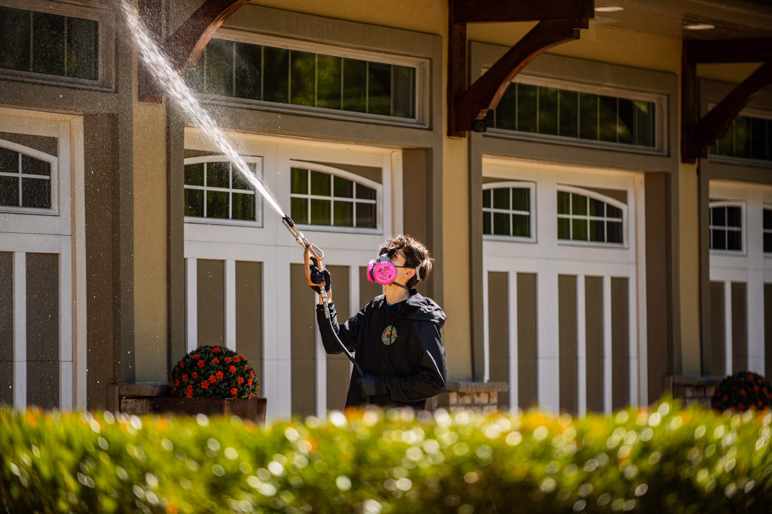 Person wearing a pink mask spraying a high-pressure water hose on garage siding outdoors.