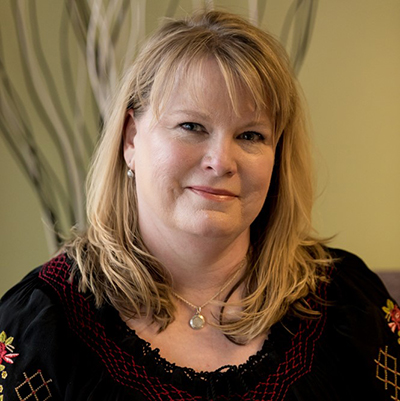 Smiling woman with blonde hair wearing a black top with red and yellow embroidery.