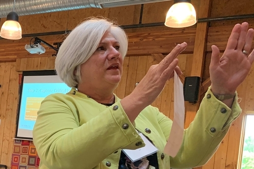 Woman with white hair in green jacket gesturing with both hands indoors near a projection screen.