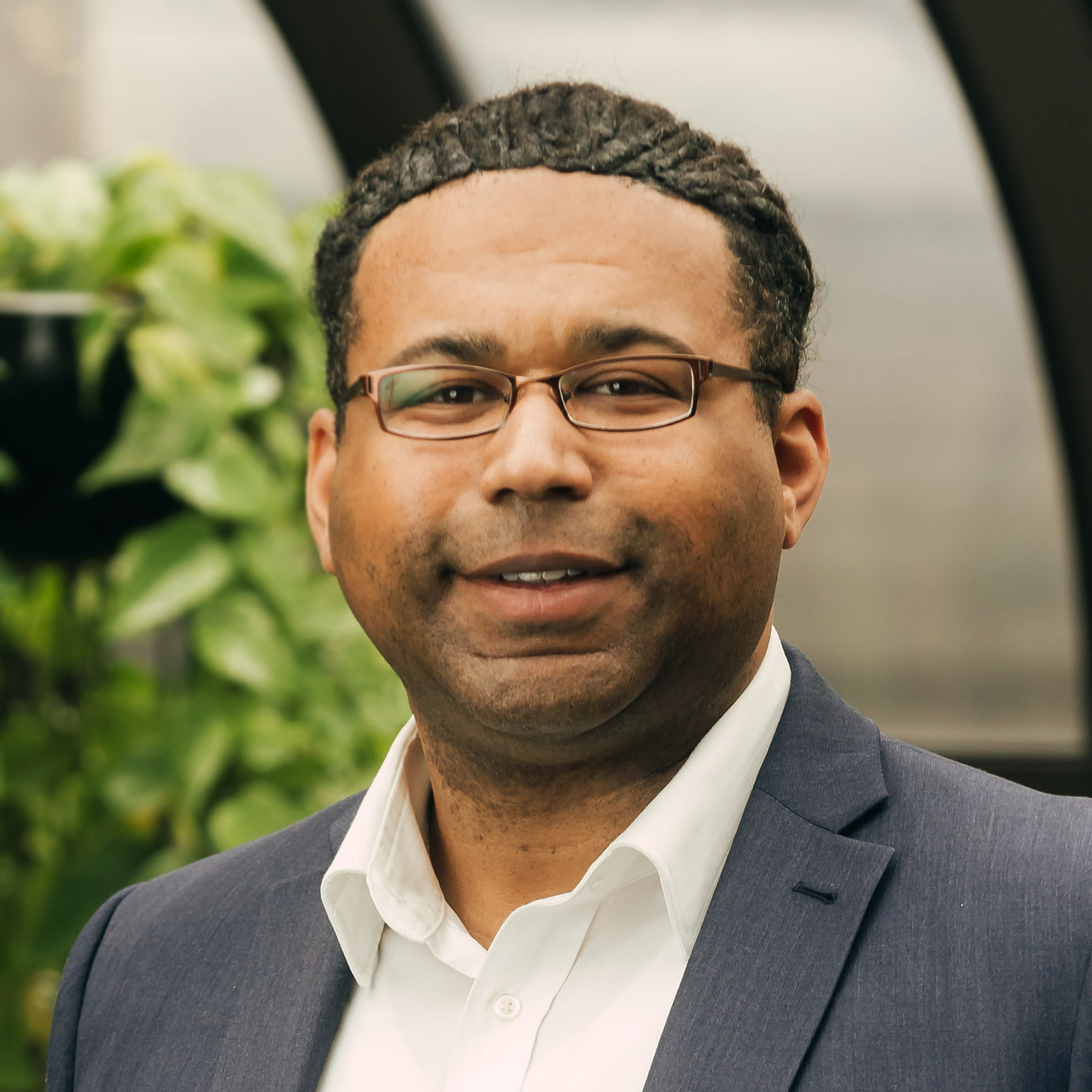 Man wearing glasses, a white shirt, and a dark blazer smiling indoors with green plants in the background.