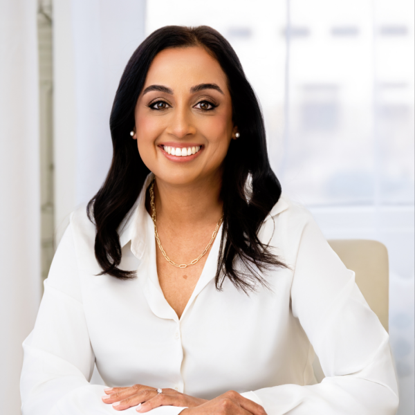 Smiling woman with dark hair wearing a white blouse and gold chain sitting at a desk.