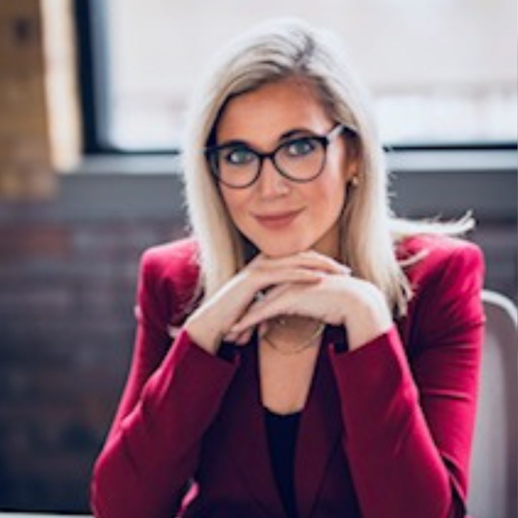 Blonde woman with glasses in a red blazer posing with hands clasped under her chin indoors.