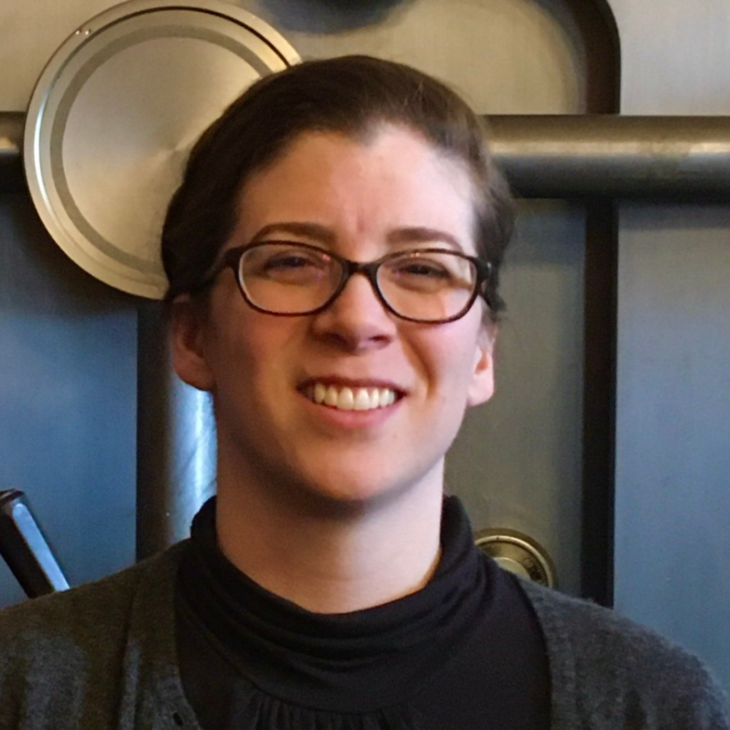 Smiling person with glasses, dark hair pulled back, wearing a black top and gray sweater.