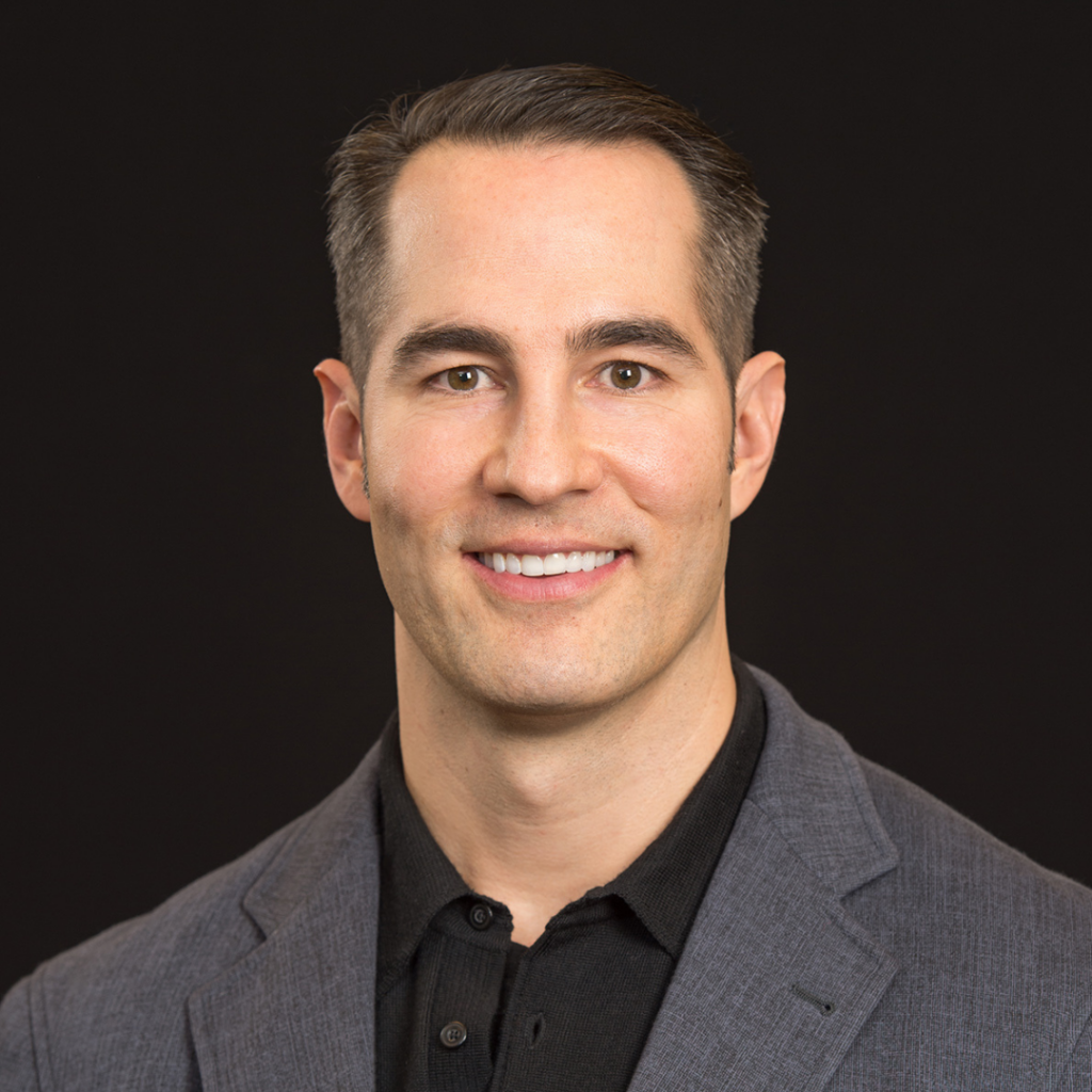 Smiling man with short dark hair wearing a gray blazer and black shirt against a black background.