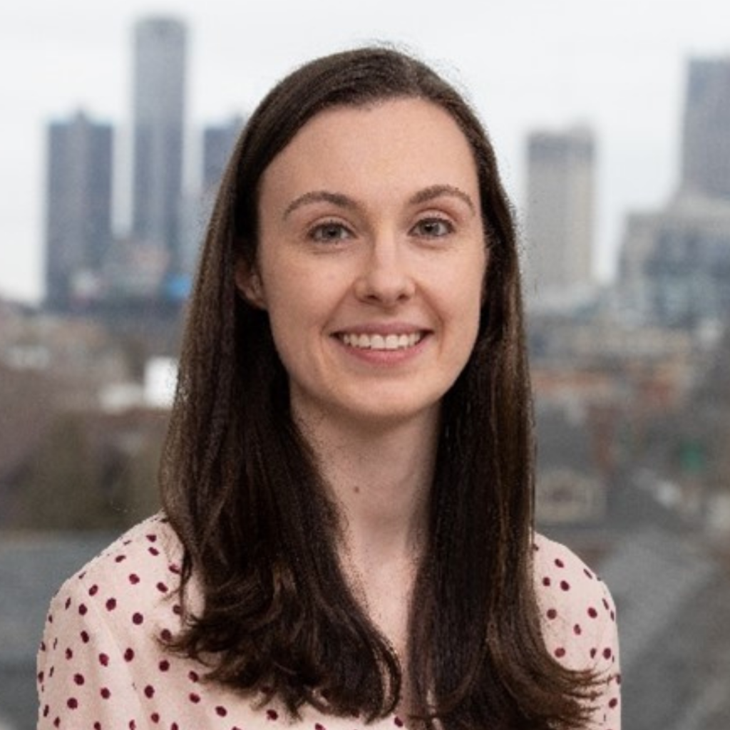 Smiling woman with long dark hair in a light shirt with red dots, city skyline blurred in background.