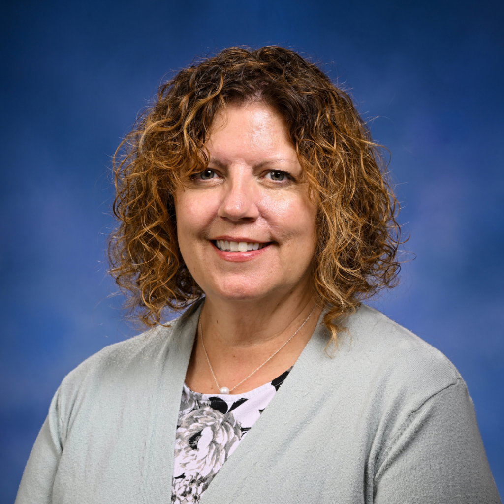 Smiling woman with curly brown hair wearing a gray cardigan and floral top on blue background.
