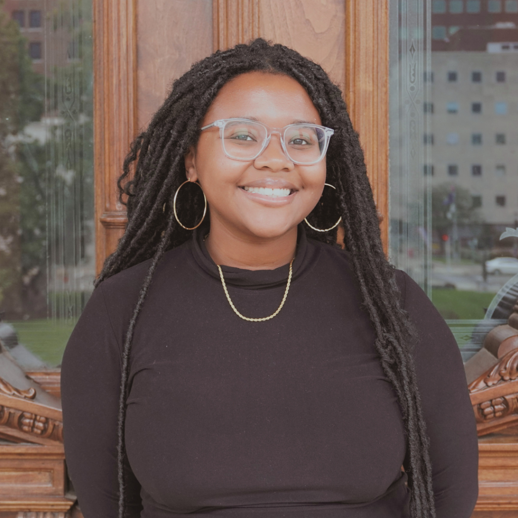 Smiling woman with long dreadlocks, clear glasses, hoop earrings, and black top in front of a wooden door.
