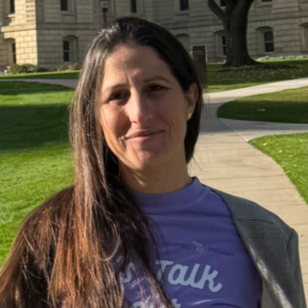 Woman with long dark hair wearing a purple shirt and gray jacket outdoors on a sunny day.