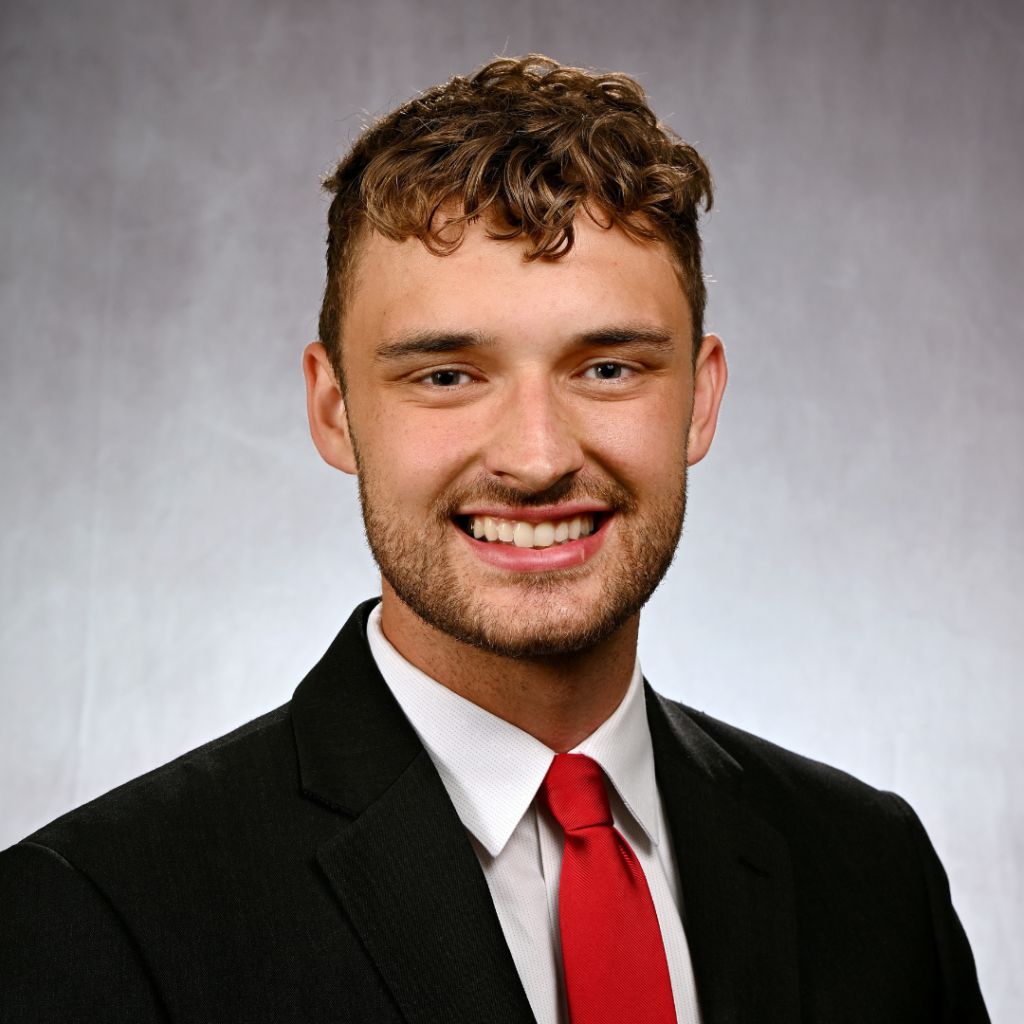 Smiling young man with curly hair wearing a black suit, white shirt, and red tie on gray background.
