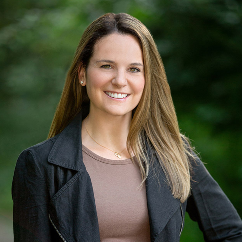 Smiling woman with long light brown hair wearing a black jacket and brown top outdoors.