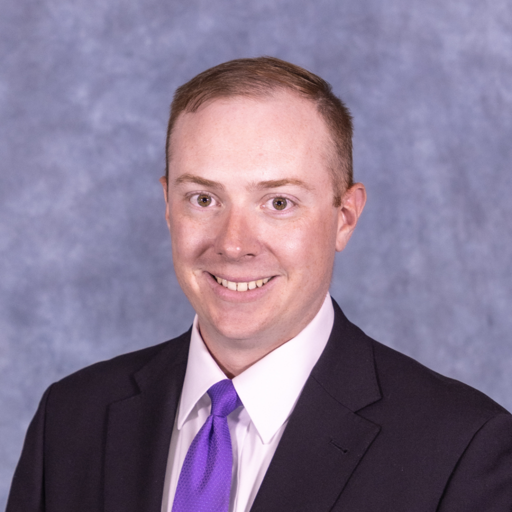 Man with light skin and brown hair wearing a black suit, white shirt, and purple tie smiling at the camera.