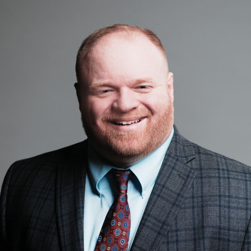 Smiling man with red beard wearing a plaid suit jacket, light blue shirt, and patterned red tie.