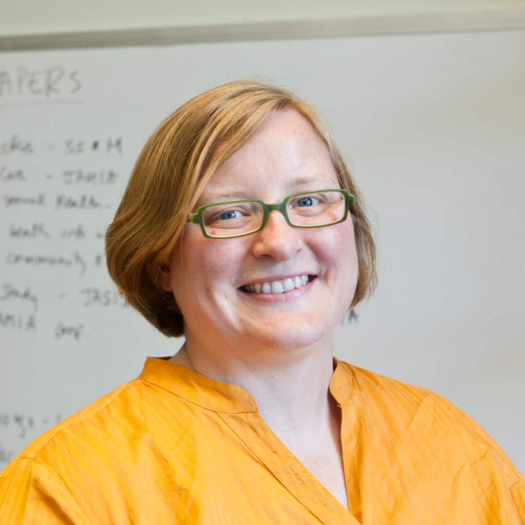 Smiling person with short blond hair and green glasses wearing a bright orange shirt.