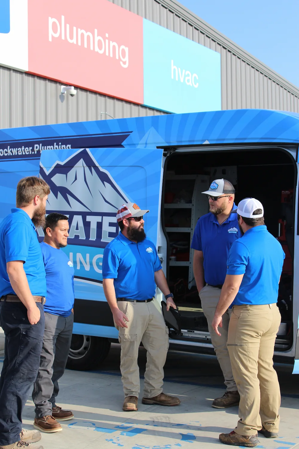 Five workers in blue shirts standing near a Rockwater Plumbing van outside a building with plumbing and hvac signs.