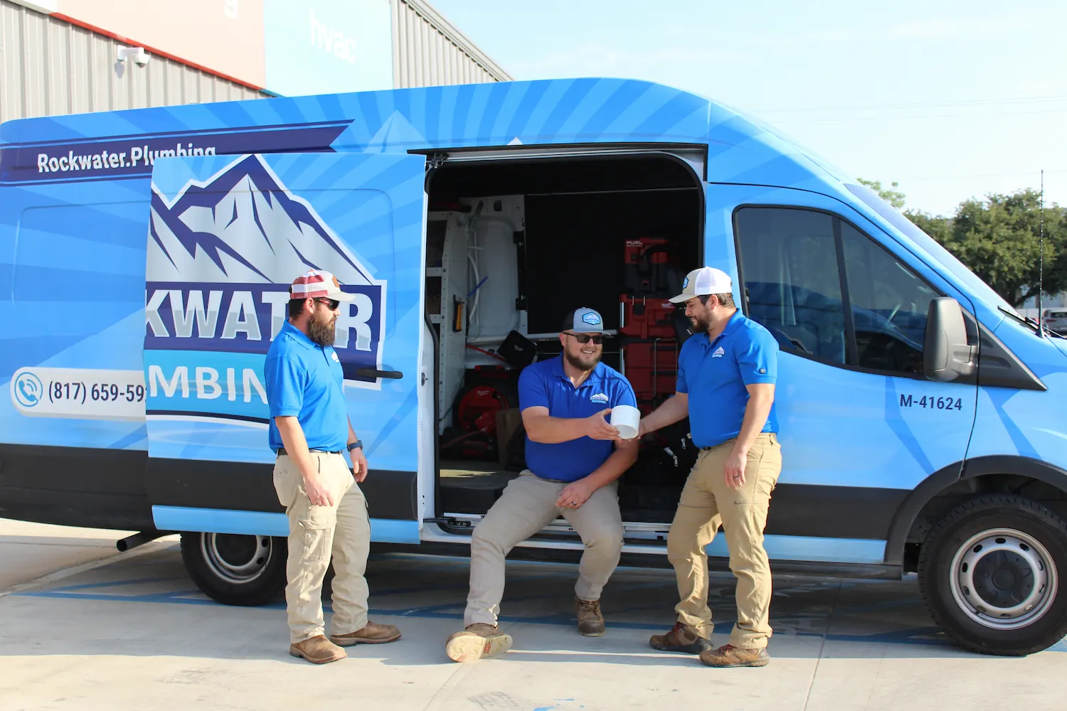 Three plumbers in blue shirts and khaki pants near a blue Rockwater Plumbing van with open door.