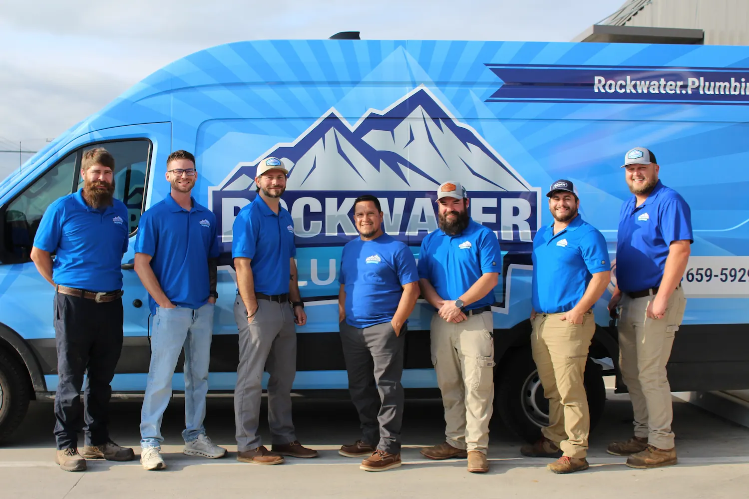 Seven men in blue shirts standing side by side in front of a blue Rockwater Plumbing van.