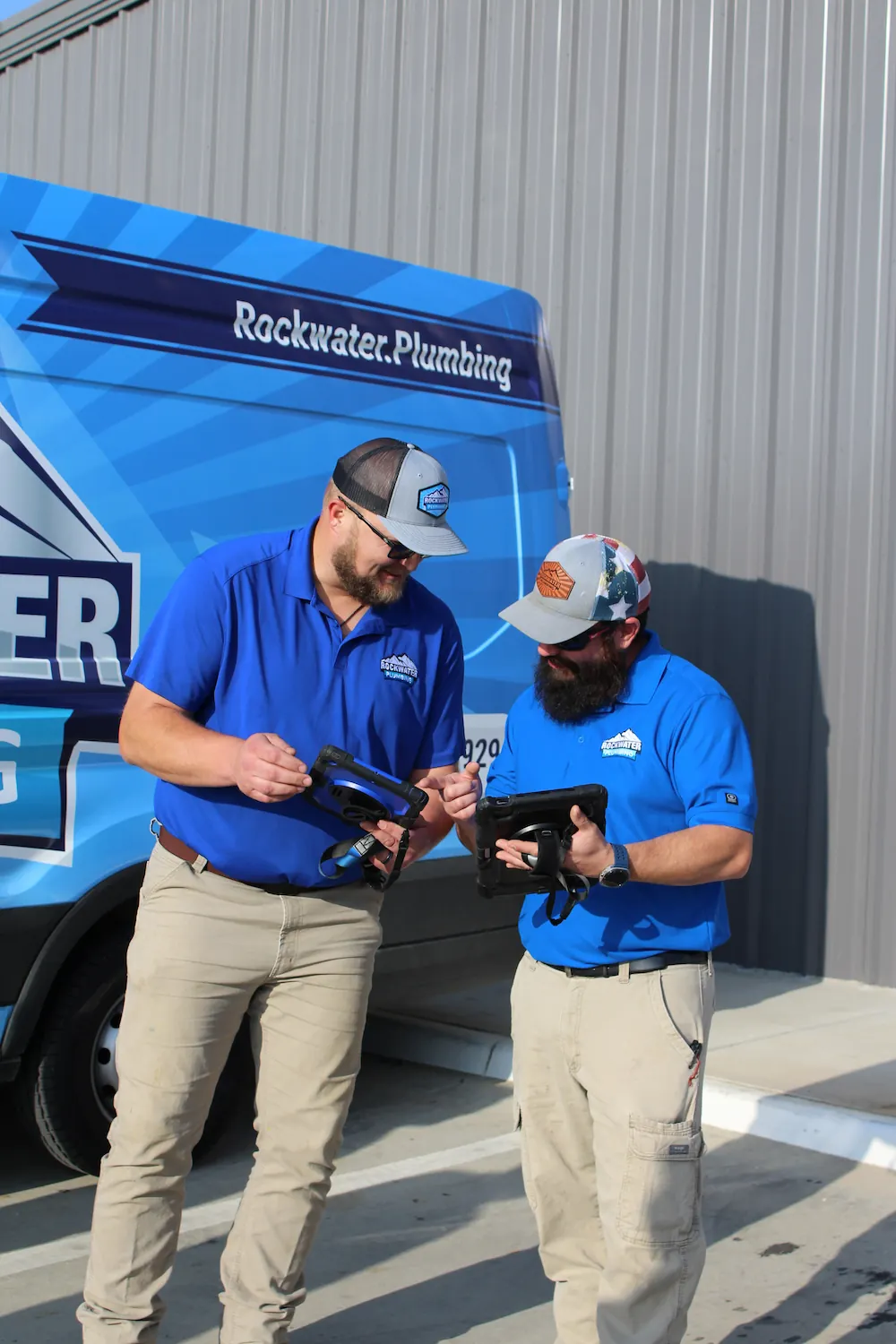 Two men in blue Rockwater Plumbing shirts stand outside a branded van using handheld devices.