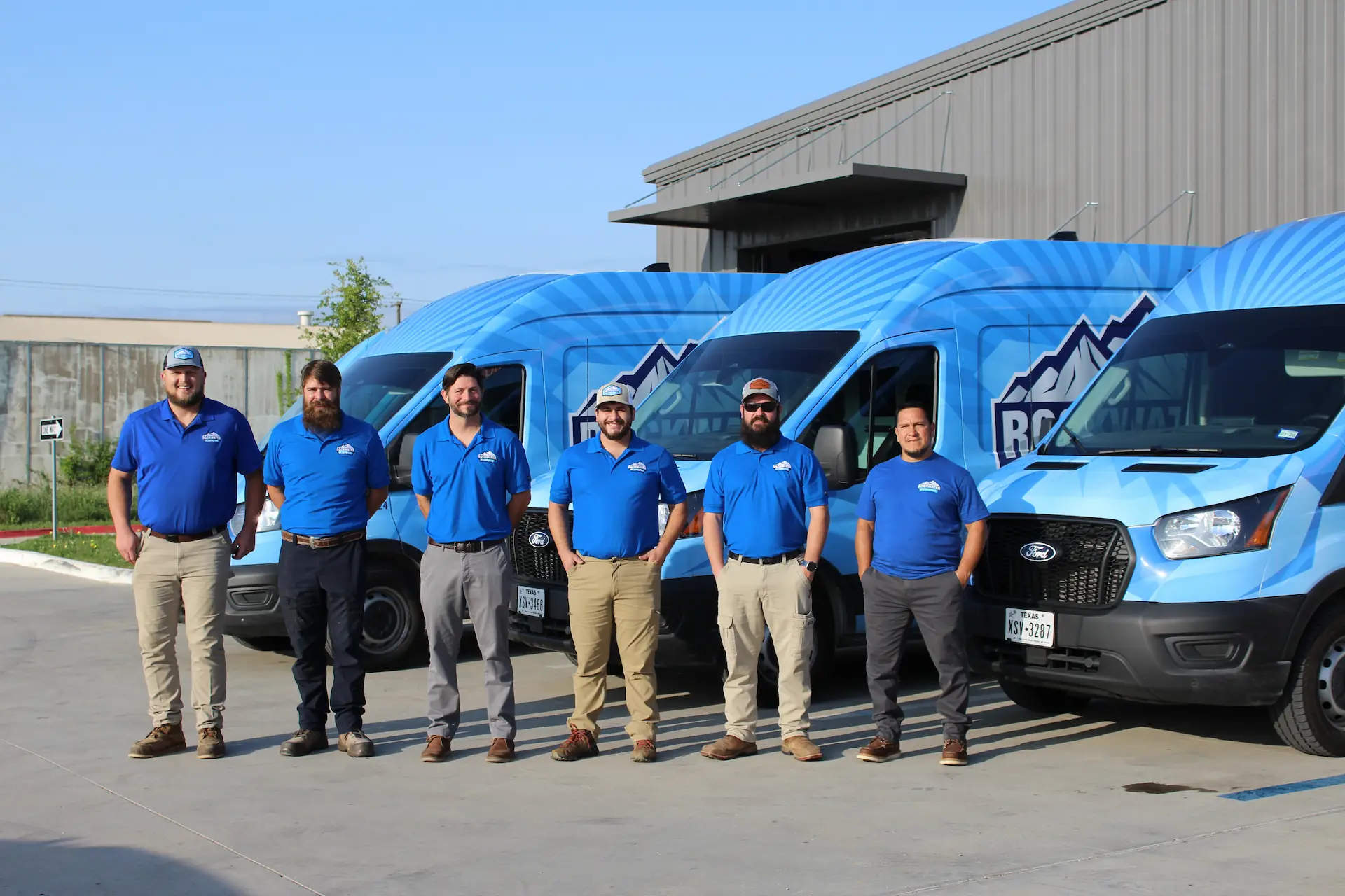 Six men in blue company shirts standing in front of blue vans with mountain logo under clear sky.
