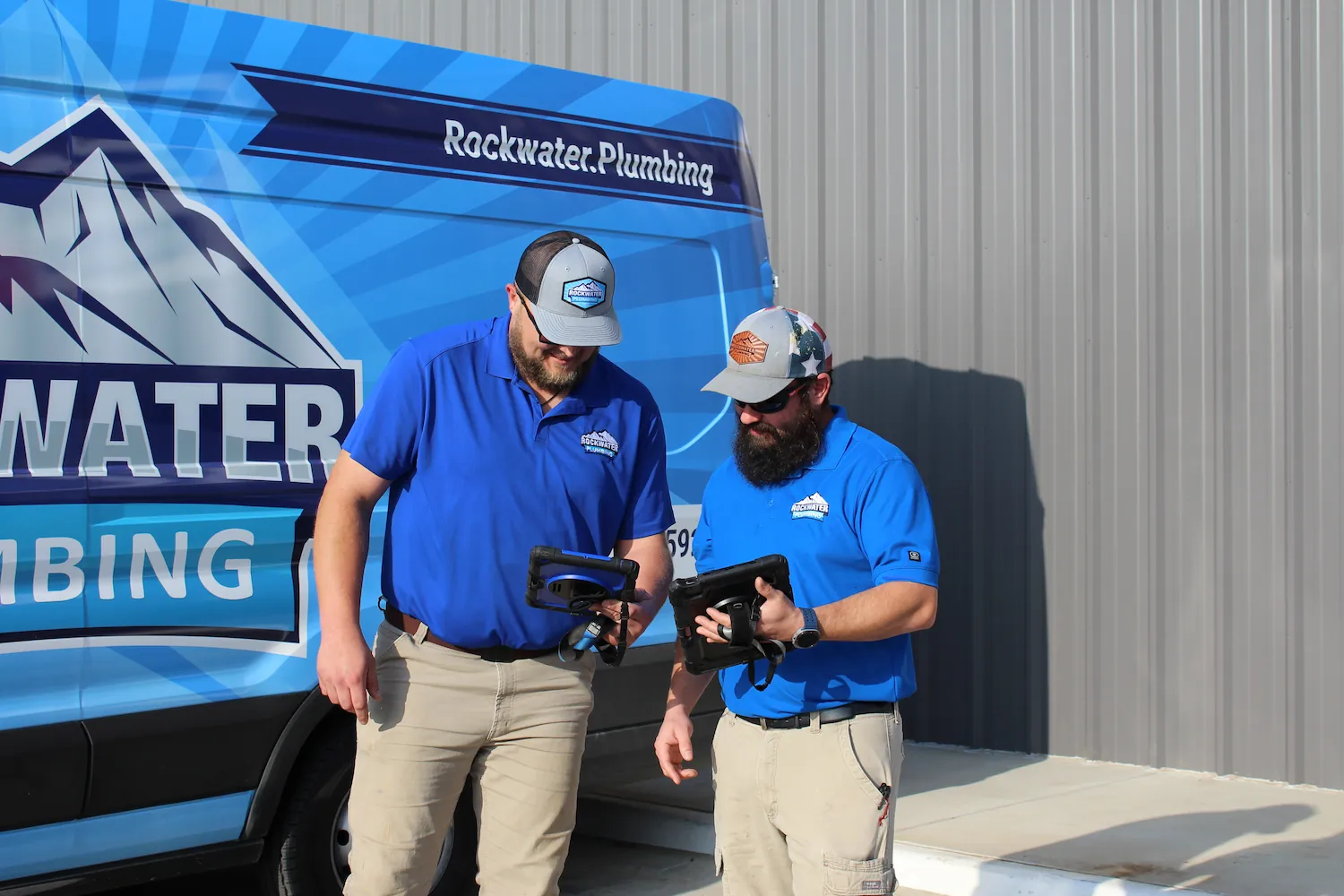 Two plumbers in blue shirts and caps looking at tablets beside a Rockwater Plumbing service van.