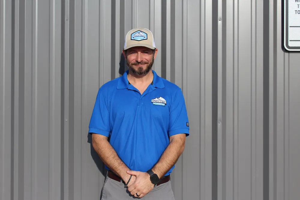 Man wearing Rockwater Plumbing uniform and cap standing against a gray metal wall smiling.