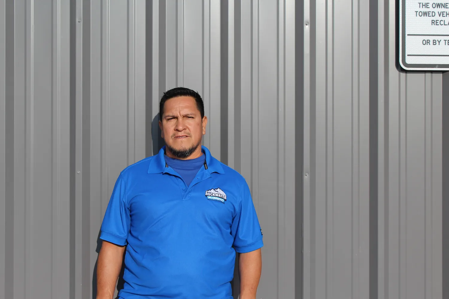 Man in blue Rockwater Plumbing polo shirt standing against a gray corrugated metal wall.