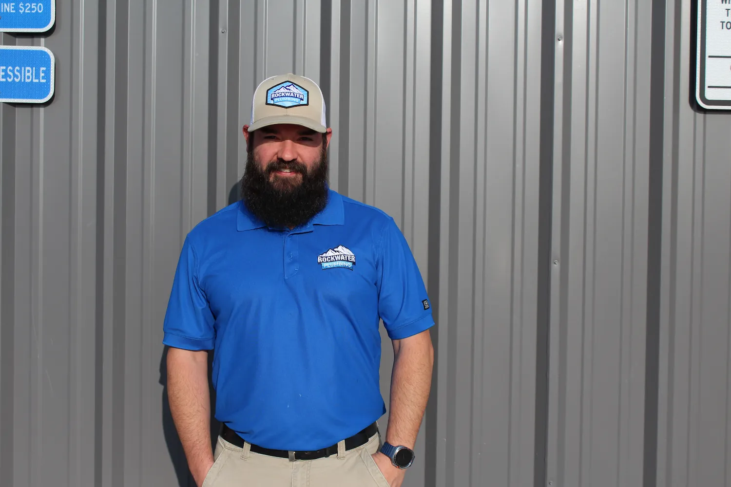 Man with beard wearing blue Rockwater Plumbing shirt and hat standing against gray metal wall.