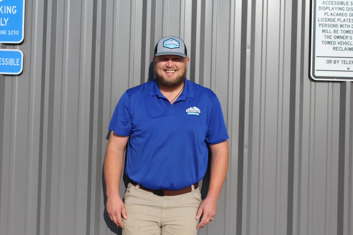 Smiling man in blue Rockwater Plumbing shirt and cap stands against gray metal wall.