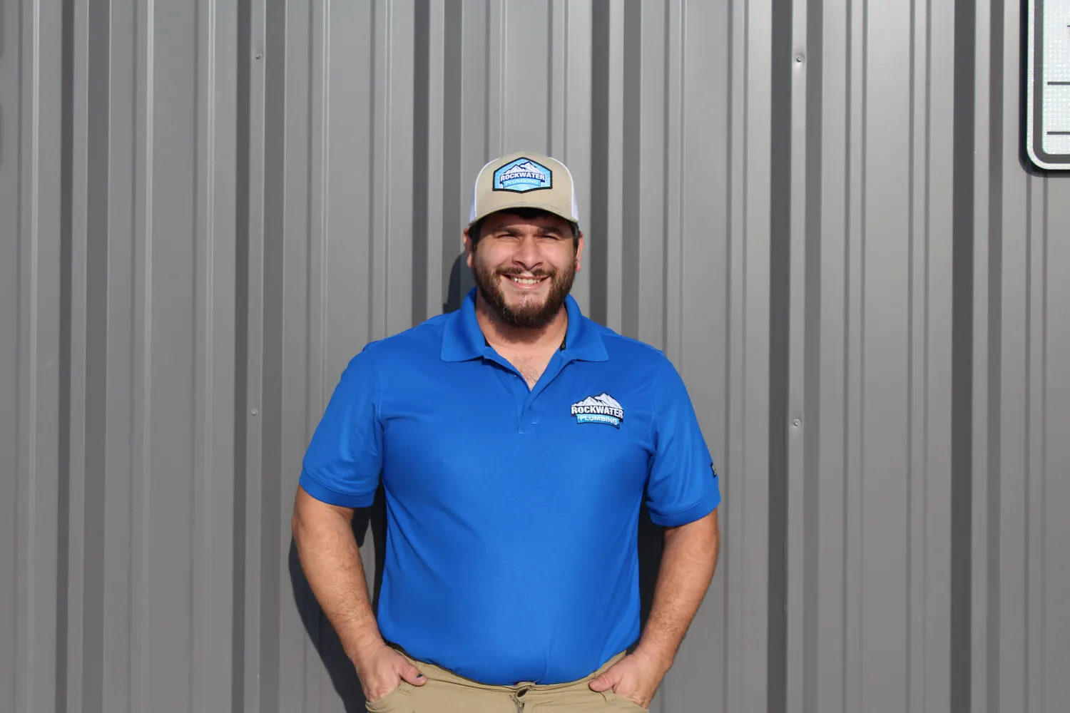 Man in a blue Rockwater Plumbing shirt and cap standing against a gray metal wall smiling.