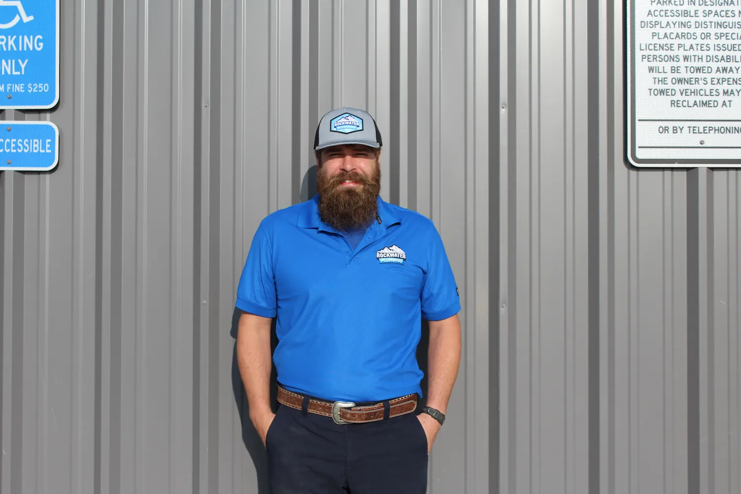 Man with beard wearing blue Rockwater Plumbing shirt and cap standing against gray corrugated wall.