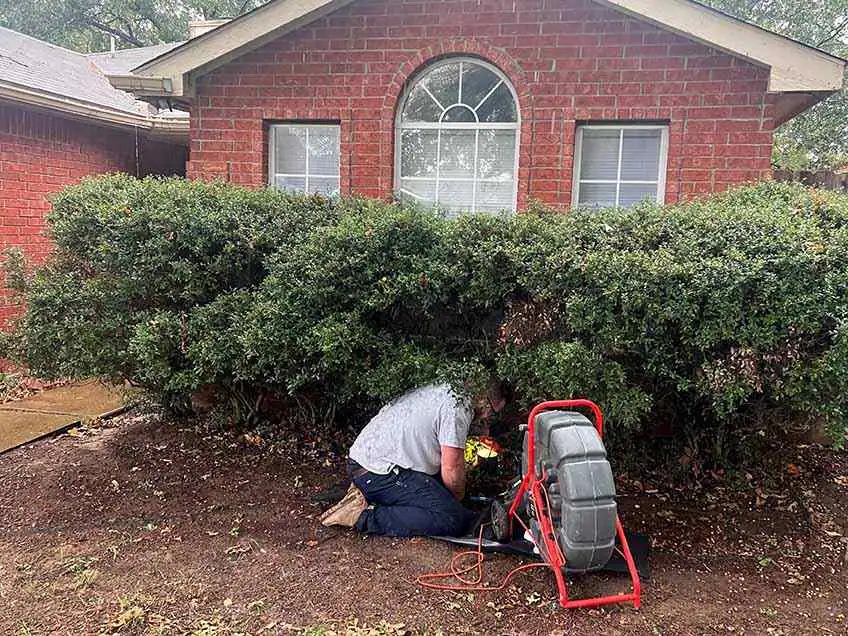 Person kneeling by a brick house using drain cleaning equipment near bushes.