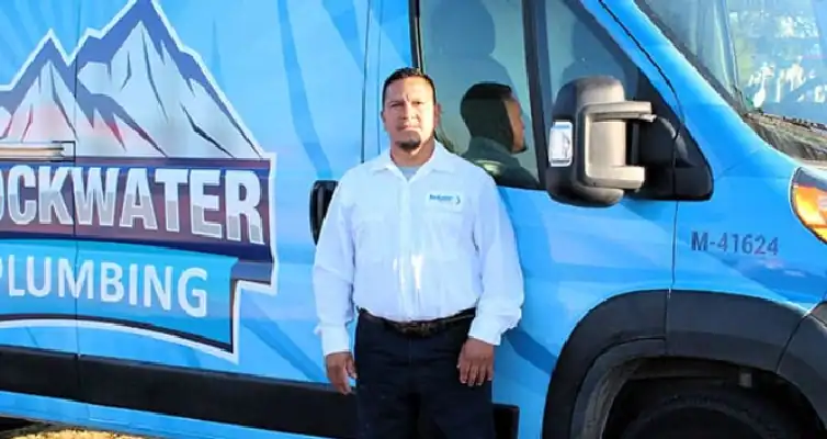 Man in white shirt standing in front of a blue Rockwater Plumbing company van with mountain logo.