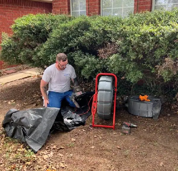 Man kneeling on soil using a plumbing inspection camera near bushes outside a brick house.