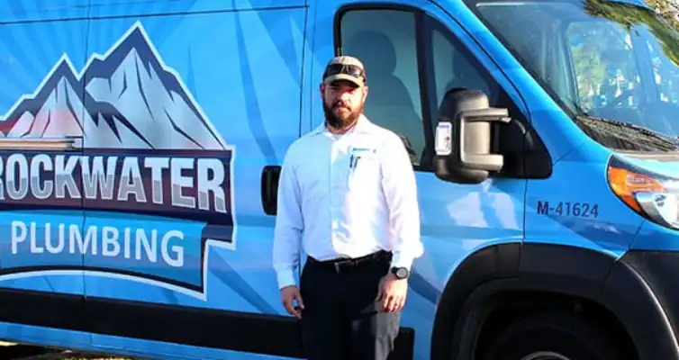 Man in white shirt and cap standing in front of a blue Rockwater Plumbing van.