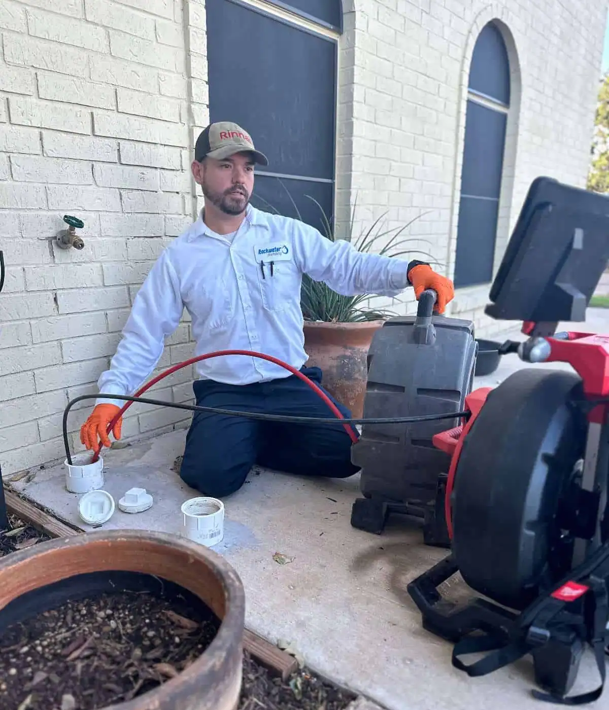 Technician in uniform using plumbing snake machine to clear outdoor drain pipe near a brick wall.
