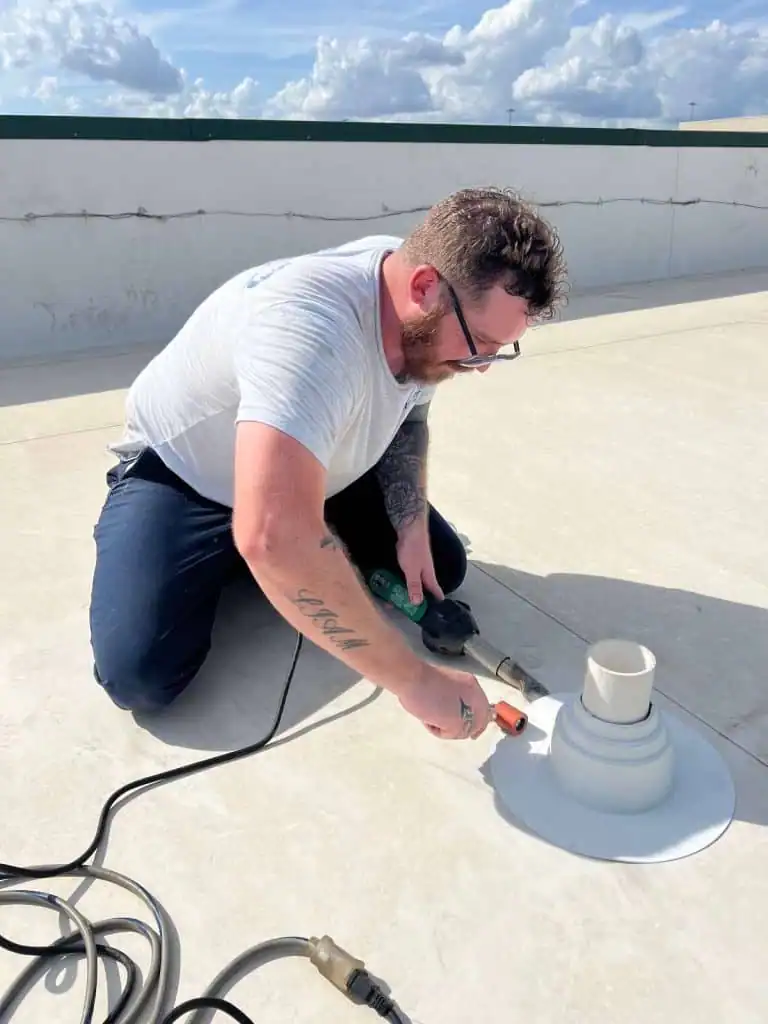 Man kneeling on a rooftop using a heat gun and roller to seal a white pipe flashing.