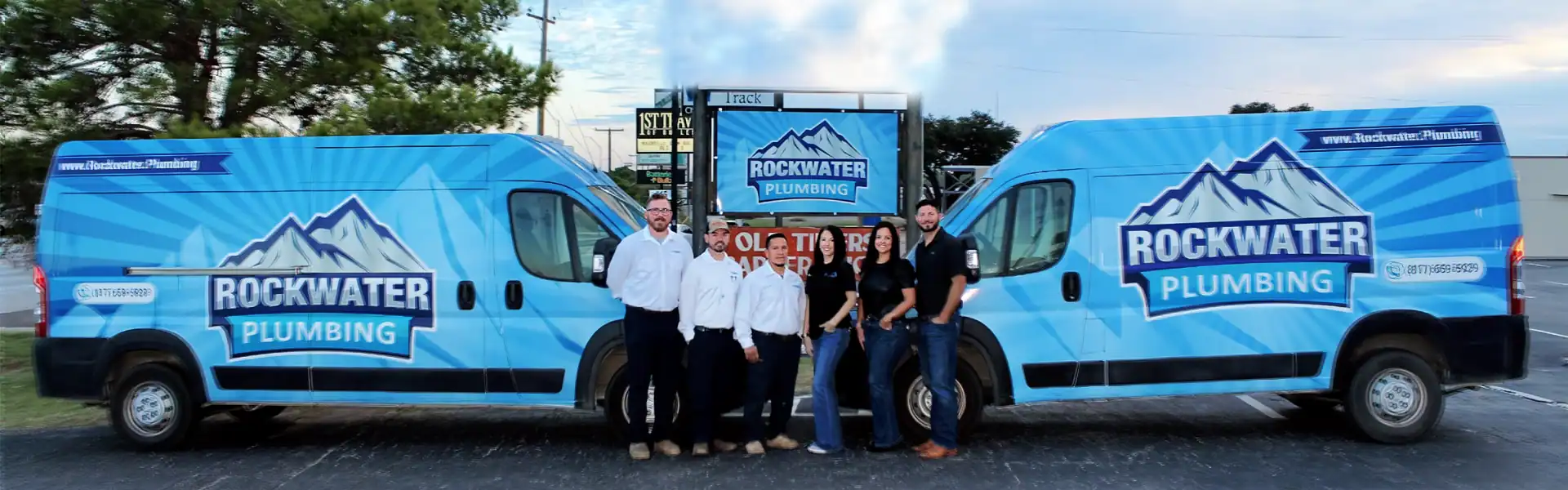 Six Rockwater Plumbing employees stand between two blue company vans with mountain logos.