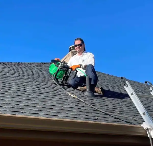 Man in sunglasses and gloves sitting on roof holding green garden hose reel next to a ladder.
