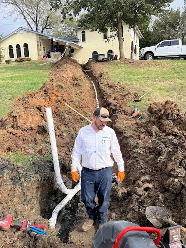 Man in work clothes standing in a trench with exposed pipes by a house and truck.