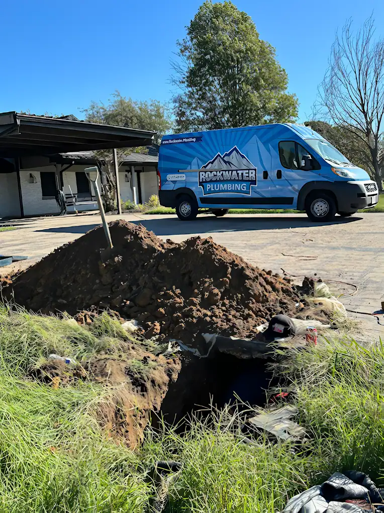 Rockwater Plumbing van parked near a large hole dug in the ground with a mound of dirt and a shovel.