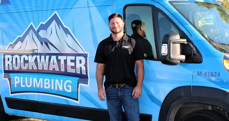 Man in black shirt and jeans standing next to a blue Rockwater Plumbing van with mountain logo.